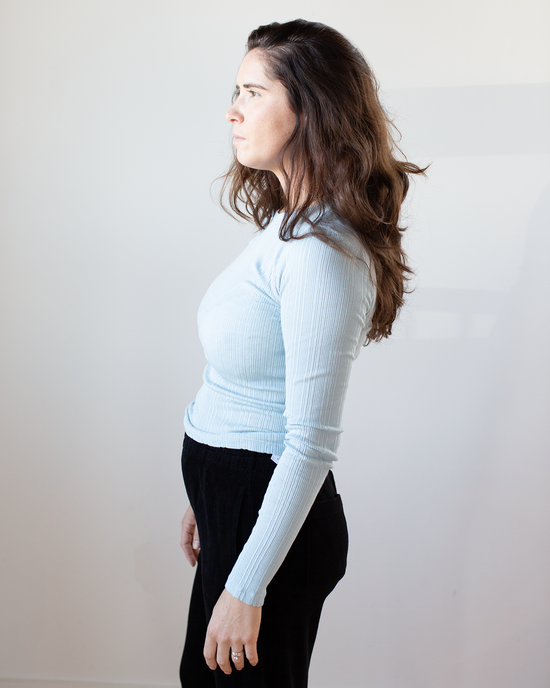 A woman with long brown hair wearing the Velvet by Graham & Spencer Mauri L/S Crew Neck Top in Ceramic and black pants stands in profile against a plain light background.