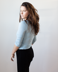 A woman with long brown hair stands sideways, looking down in a slim fit Nima L/S Boatneck Top in Ceramic by Velvet by Graham & Spencer and black pants, set against a plain light background.