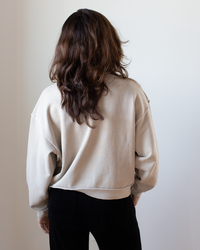 A person with wavy brown hair faces away, wearing the NSF Kenya Oversize FT Crew Sweatshirt in Faded Taupe with black pants, set against a plain white background.