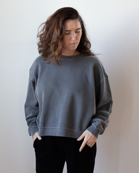 A person with wavy brown hair wears the NSF Kenya Oversize FT Crew Sweatshirt in Faded Black and black pants, standing against a plain white background with hands in pockets, looking down.