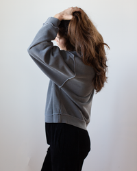 A woman with long brown hair, dressed in the NSF Kenya Oversize FT Crew Sweatshirt in Faded Black and black pants, stands sideways against a plain light background, lifting her hair with one hand while her face is partially turned away.