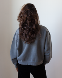 A person with long, wavy brown hair stands with their back to the camera, wearing the NSF Kenya Oversize FT Crew Sweatshirt in Faded Black and black pants against a plain light background.