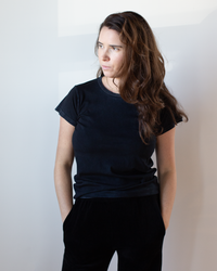 A woman with long brown hair wears a Free City Canyonkid Circa1972 Supervintage Tee in Superblack Powder and black pants, standing with her hands in her pockets and looking thoughtfully to her left against a plain white background.