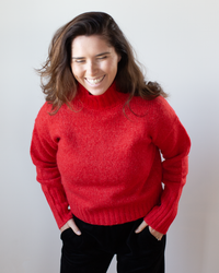 A person with long brown hair smiles, eyes closed, wearing Hartford's Myac Turtleneck Sweater in Lipstick—a cozy wool alpaca blend pullover—and black pants, standing against a plain light background with hands in pockets.