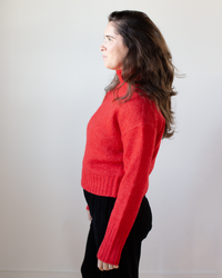 A woman with long brown hair, dressed in the Hartford Myac Turtleneck Sweater in Lipstick and black pants, stands in profile against a plain white background.
