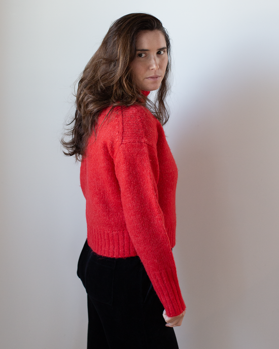 A person with long brown hair, wearing the Hartford Myac Turtleneck Sweater in Lipstick and black pants, stands sideways and looks at the camera against a plain white background.