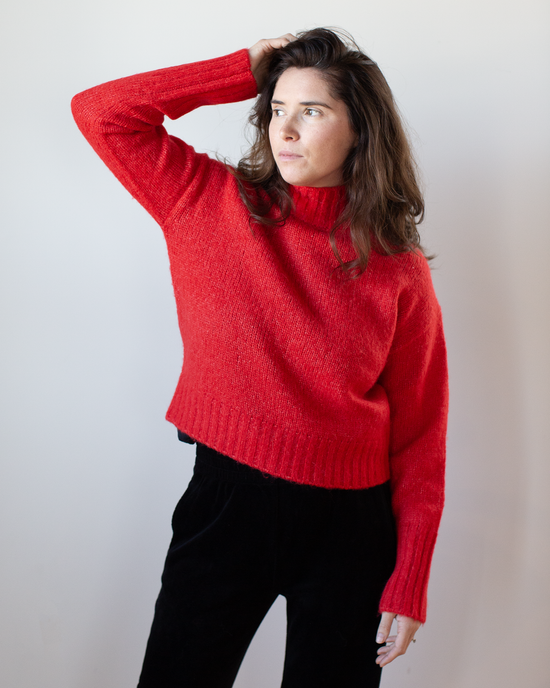 A woman with long brown hair wears the Hartford Myac Turtleneck Sweater in Lipstick and black pants, standing against a plain light background while touching her head and looking to the side.