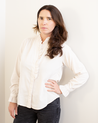 A woman with long brown hair models the Hartford Carmela Ruffle Collar Shirt in Stone, paired with dark pants. She stands against a plain light wall, looking at the camera with a neutral expression and one hand on her hip.
