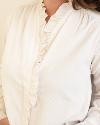 A person wears the Hartford Carmela Ruffle Collar Shirt in Stone, featuring a light hue, long sleeves, ruffle collar, delicate front ruffles, subtle striped texture, and a relaxed fit. The person's face is partially out of frame.