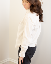A woman with long brown hair stands sideways in a well-lit corner, wearing the Hartford Carmela Ruffle Collar Shirt in Stone and dark jeans. She looks down softly, her outfit creating a relaxed silhouette.