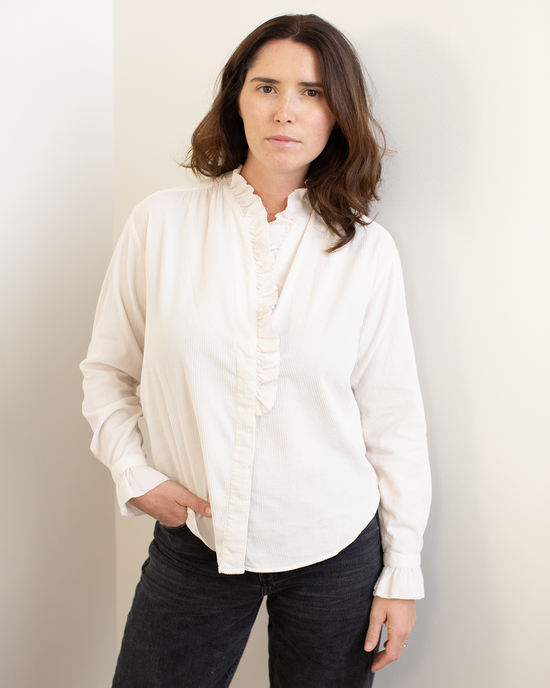 A woman with wavy brown hair stands against a light wall, wearing the Hartford Carmela Ruffle Collar Shirt in Stone and black jeans, one hand in her pocket, her neutral expression and pose creating a relaxed silhouette.