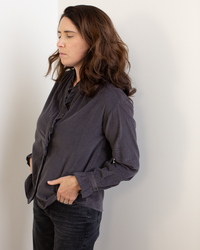 A woman stands against a white wall with her hands in her pockets, wearing the Hartford Carmela Ruffle Collar Shirt in Anthracite and dark jeans, her long brown hair down and her gaze directed to the side with a neutral expression.