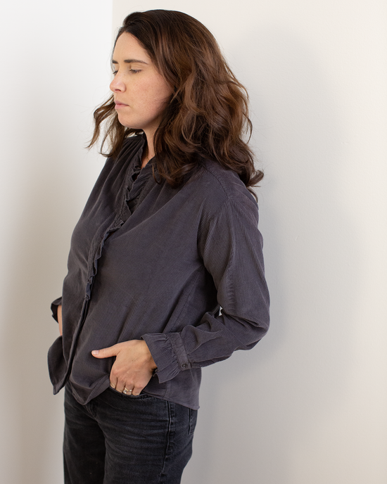 A woman stands against a white wall with her hands in her pockets, wearing the Hartford Carmela Ruffle Collar Shirt in Anthracite and dark jeans, her long brown hair down and her gaze directed to the side with a neutral expression.