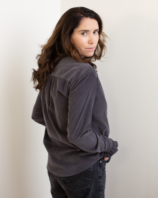 A woman with long brown hair stands sideways against a light background, looking over her shoulder at the camera while wearing Hartford's Carmela Ruffle Collar Shirt in Anthracite and black jeans.