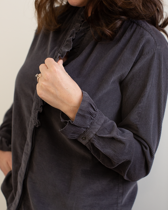A person with wavy brown hair wears the Hartford Carmela Ruffle Collar Shirt in Anthracite, holding the collar to reveal a ring. The background is plain and light.