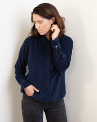 A woman with shoulder-length brown hair wears the Hartford Carmela Ruffle Collar Shirt in Midnight and black jeans, standing against a light wall, looking down with one hand on her ear and the other in her pocket.