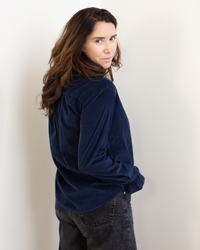 A person with long, wavy brown hair wears the Hartford Carmela Ruffle Collar Shirt in Midnight, paired with dark jeans, standing against a plain light background and glancing over their shoulder at the camera.