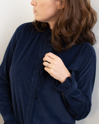 A woman with wavy brown hair wears the Hartford Carmela Ruffle Collar Shirt in Midnight and a ring on her left hand, held near her collar. She looks to the side, away from the camera.