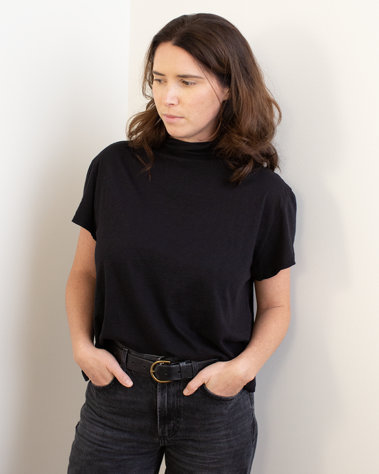 A woman with wavy brown hair, wearing the Savi S/S Mock Neck Crop Top in Black by Velvet by Graham & Spencer and black jeans with a belt, stands against a white wall with her hands in her pockets, looking to the side.