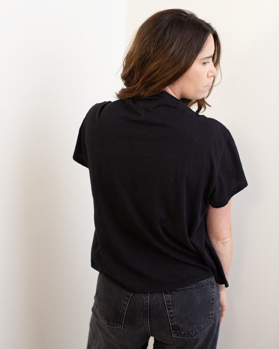 A woman with brown hair wears the Velvet by Graham & Spencer Savi S/S Mock Neck Crop Top in Black and dark jeans, standing with her back to the camera and turning her head slightly to the right in a neutral, light-colored room.