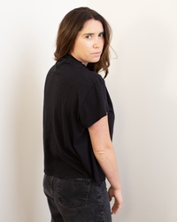 A woman with long brown hair wears the Savi S/S Mock Neck Crop Top in Black by Velvet by Graham & Spencer and dark jeans, standing sideways with a neutral expression, looking over her shoulder at the camera against a plain light background.