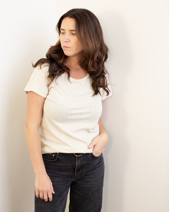 A woman with long brown hair, wearing the Velvet by Graham & Spencer Odelia Scoop Neck Tee in Cookie and black jeans, stands against a white wall looking to her left with a neutral expression.
