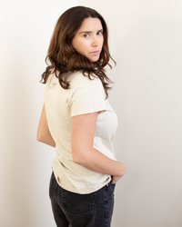 A woman with long brown hair, wearing the Odelia Scoop Neck Tee in Cookie by Velvet by Graham & Spencer and dark jeans, stands side-on with her hands in her pockets, glancing over her shoulder with a neutral expression.
