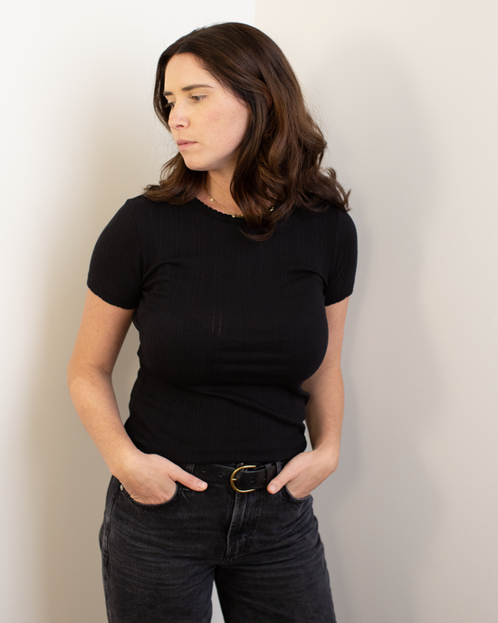 A woman with shoulder-length brown hair wears the Enati S/S Crew Neck Top in Black by Velvet by Graham & Spencer and dark jeans, standing against a white wall with her hands in her pockets, looking to the side with a neutral expression.