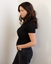 A woman with wavy brown hair, wearing the Enati S/S Crew Neck Top in Black by Velvet by Graham & Spencer and black jeans, stands in profile against a white wall with her hands in her pockets, looking downward.