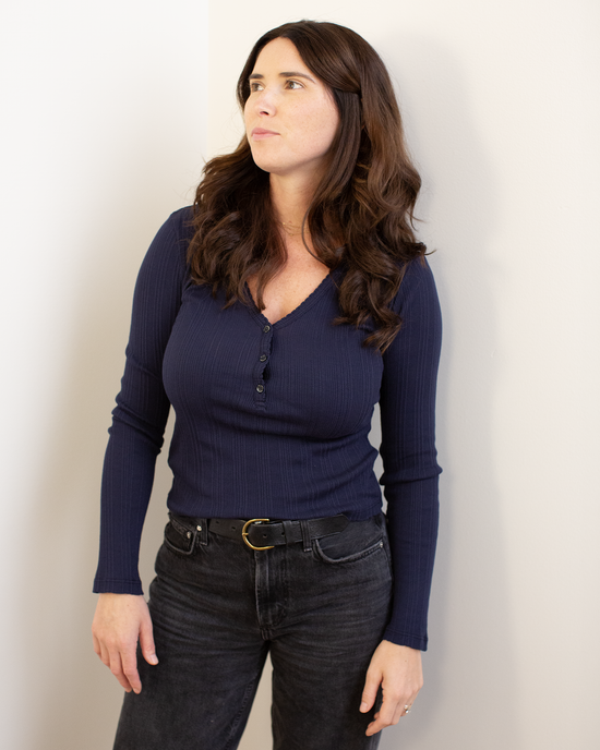 A woman with long brown hair, wearing the Velvet by Graham & Spencer Winona L/S Button Top in Navy and black jeans, stands against a light-colored wall, looking slightly to the side.