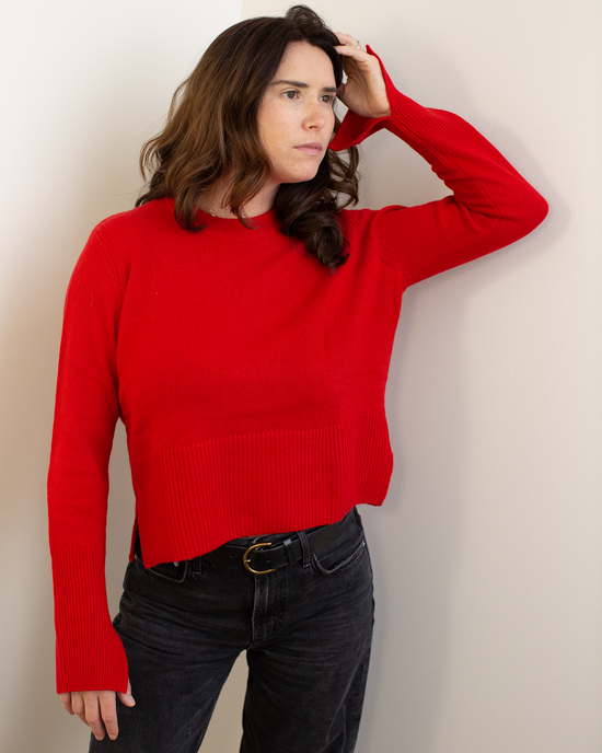 A woman with wavy brown hair wears the Autumn Cashmere Cropped Boxy Crew W/ Slits in Blazer, paired with black jeans, standing against a light wall and looking thoughtfully to the side with one hand touching her head.