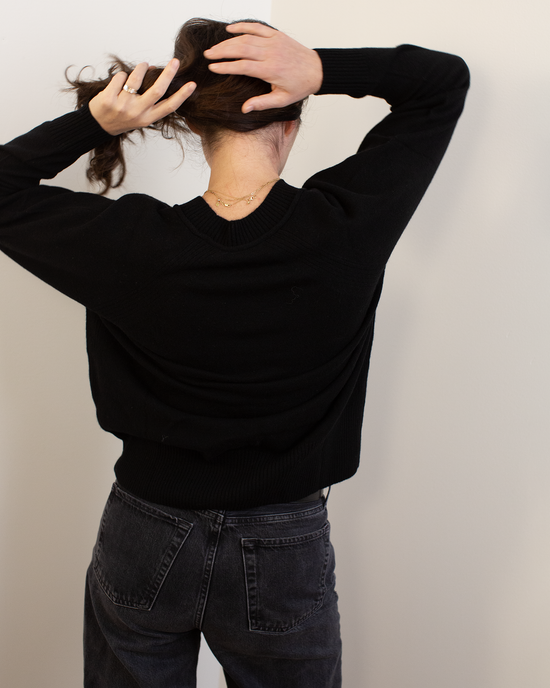 A person with dark hair, wearing Autumn Cashmere's Relaxed Fit Raglan Crew with Rib Detail in Black and dark jeans, stands facing a white wall with their back to the camera, both hands raised as they gather their hair.