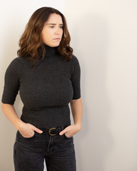 A woman with wavy brown hair wears the Autumn Cashmere Cropped Elbow Sleeve Rib Turtleneck in Pepper and black jeans, standing with her hands in her pockets and looking to the side against a plain light-colored wall.