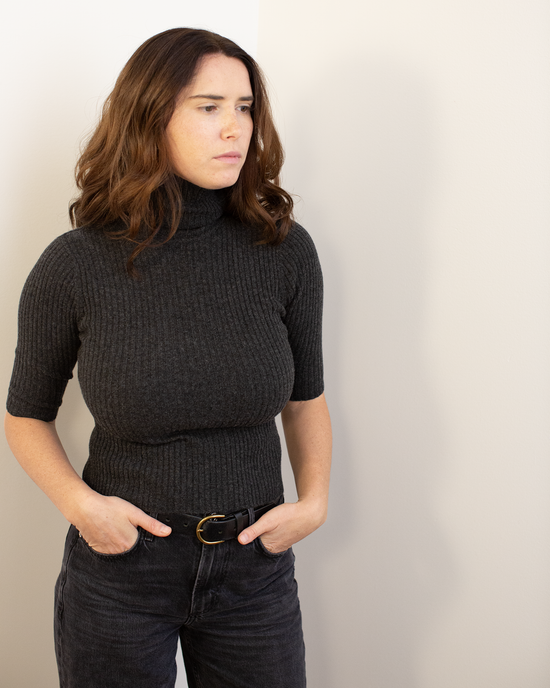 A woman with wavy brown hair wears the Autumn Cashmere Cropped Elbow Sleeve Rib Turtleneck in Pepper and black jeans, standing with her hands in her pockets and looking to the side against a plain light-colored wall.