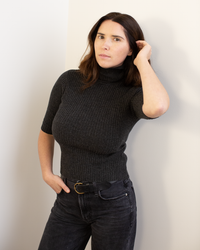A woman with long brown hair stands against a light wall, looking at the camera with a neutral expression, one hand touching her hair. She wears the Autumn Cashmere Cropped Elbow Sleeve Rib Turtleneck in Pepper and black jeans.