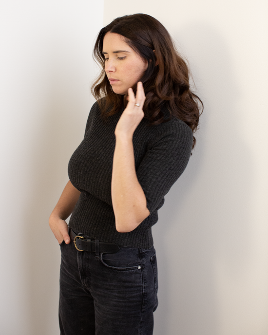 A woman with wavy brown hair stands against a white wall, wearing the Autumn Cashmere Cropped Elbow Sleeve Rib Turtleneck in Pepper. One hand touches her neck, the other in her pocket, as she looks downward with a calm expression.