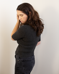 A woman with long brown hair wears the Autumn Cashmere Cropped Elbow Sleeve Rib Turtleneck in Pepper with dark jeans, standing against a white wall and looking down while touching the side of her face.