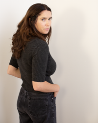 A woman with long brown hair wears the Autumn Cashmere Cropped Elbow Sleeve Rib Turtleneck in Pepper and black jeans, standing sideways with her hand in her pocket, looking at the camera against a plain light background.