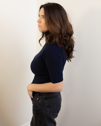 A woman with long brown hair stands in profile against a light wall, wearing the Autumn Cashmere Cropped Elbow Sleeve Rib Turtleneck in Navy with black jeans, her hand in her pocket as she looks ahead.