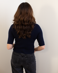 A person with long, wavy brown hair stands facing a white wall, wearing the Autumn Cashmere Cropped Elbow Sleeve Rib Turtleneck in Navy and high-waisted black jeans. Their arms are relaxed at their sides.