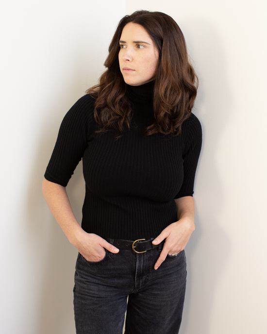 A woman with wavy brown hair models the Autumn Cashmere Cropped Elbow Sleeve Rib Turtleneck in Black with dark jeans, standing against a light wall, looking to the side with one hand in her pocket and the other on her hip.