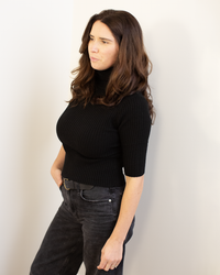 A woman with long brown hair wears the Autumn Cashmere Cropped Elbow Sleeve Rib Turtleneck in Black, paired with dark jeans, as she stands against a white wall looking to the side with a neutral expression.