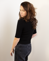A woman with long brown hair, wearing the Autumn Cashmere Cropped Elbow Sleeve Rib Turtleneck in Black and dark jeans, stands against a light backdrop, looking over her shoulder with a neutral expression.