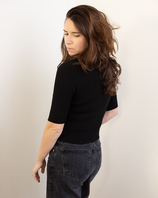 A woman with long brown hair, wearing the Autumn Cashmere Cropped Elbow Sleeve Rib Turtleneck in Black and dark jeans, stands against a light backdrop, looking over her shoulder with a neutral expression.