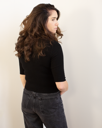 A woman with long, wavy brown hair, turned to the side with her back mostly to the camera, wears the Autumn Cashmere Cropped Elbow Sleeve Rib Turtleneck in Black and high-waisted dark jeans against a plain light background.