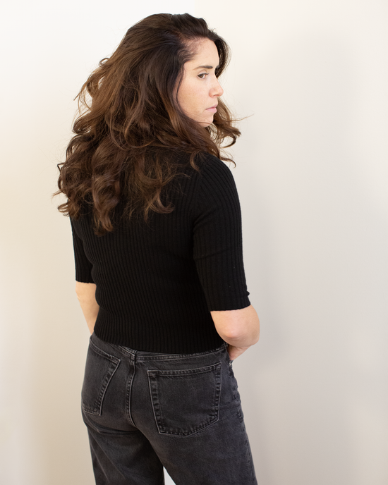 A woman with long, wavy brown hair, turned to the side with her back mostly to the camera, wears the Autumn Cashmere Cropped Elbow Sleeve Rib Turtleneck in Black and high-waisted dark jeans against a plain light background.