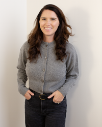 A woman with long brown hair smiles with her hands in her pockets, wearing black jeans and the Autumn Cashmere Cropped Cardigan in Cement, standing against a white wall.