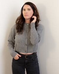 A woman with long brown hair wears the Autumn Cashmere Cropped Cardigan in Cement and black belted jeans, standing against a light wall and looking at the camera with a neutral expression.