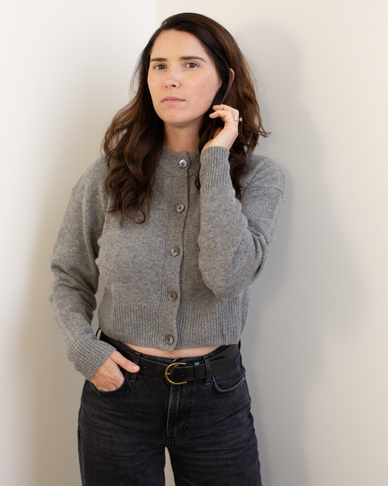 A woman with long brown hair wears the Autumn Cashmere Cropped Cardigan in Cement and black belted jeans, standing against a light wall and looking at the camera with a neutral expression.