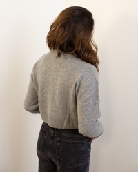 A person with wavy brown hair, facing away from the camera, wears the Autumn Cashmere Cropped Cardigan in Cement with black jeans against a plain light backdrop.